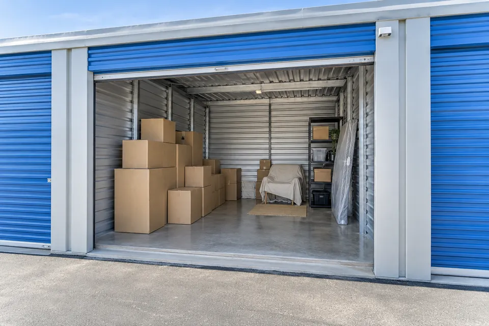 Illustrative campaign image: clean self-storage unit with neatly stacked boxes and a covered chair.